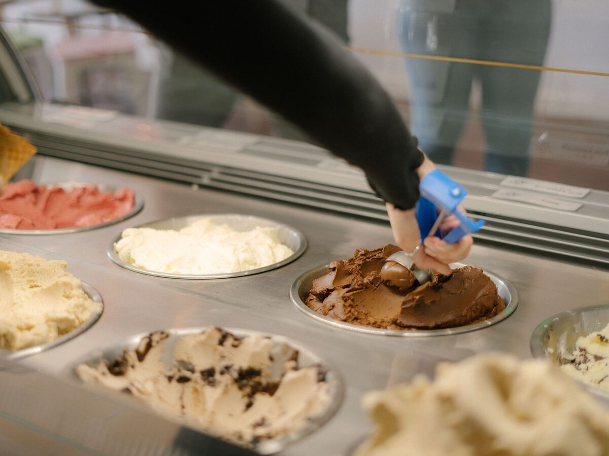 Mano porcionando helado de chocolate artesanal con una cuchara de resorte, sobre las cubetas de una heladería
