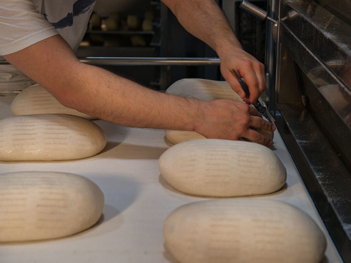 Manos de panadero greñando hogazas de masa madre con una cuchilla, junto a la boca del horno